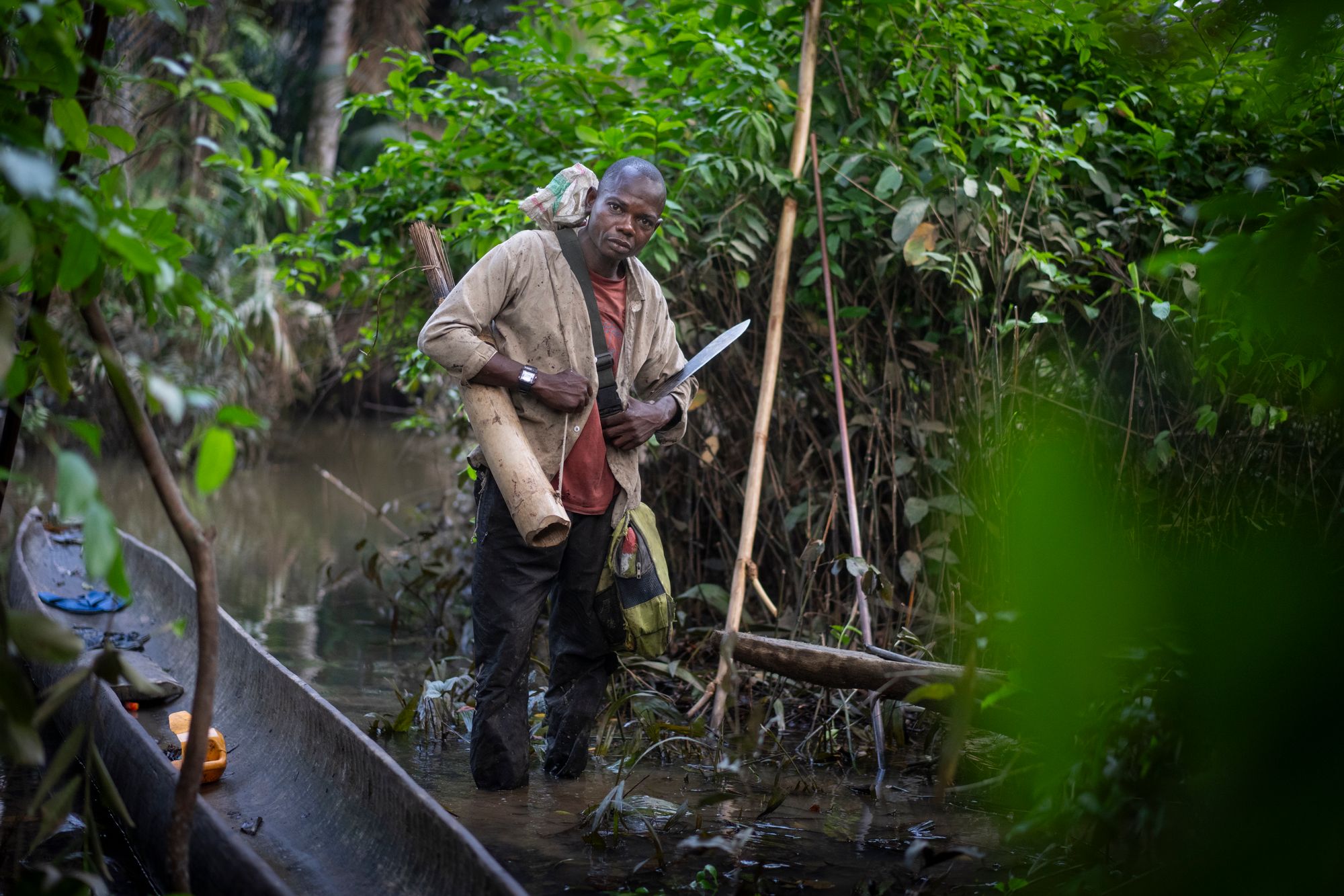 Man standing near dugout canoe in dense tropical forest, holding machete
